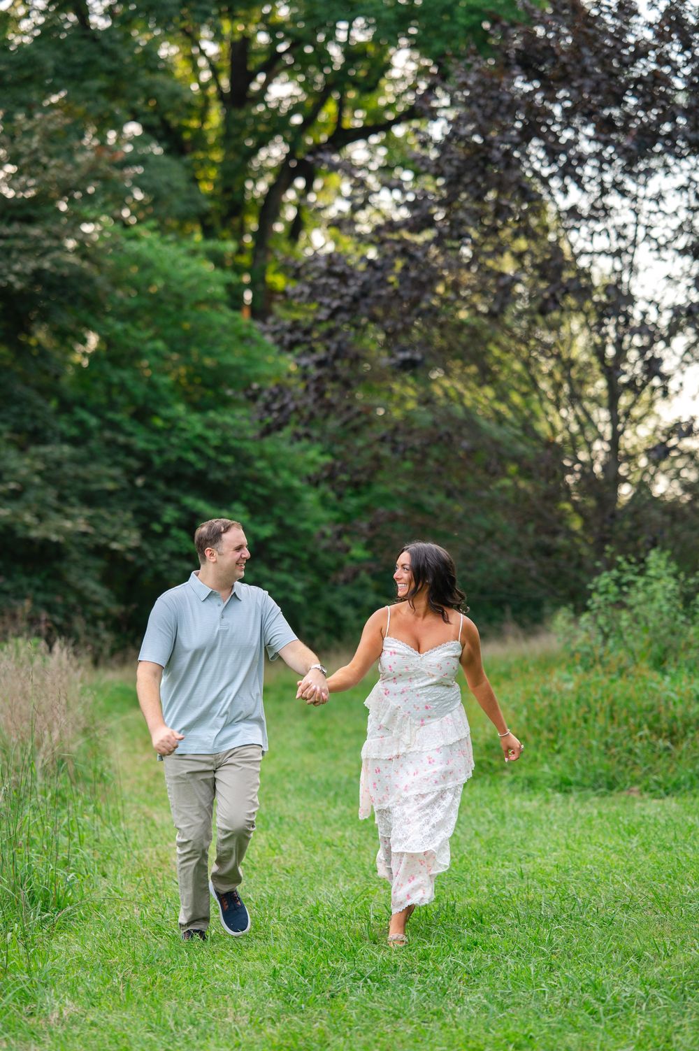 Couple holding hands walking through lush green meadow, woman in white dress and man in blue polo shirt.