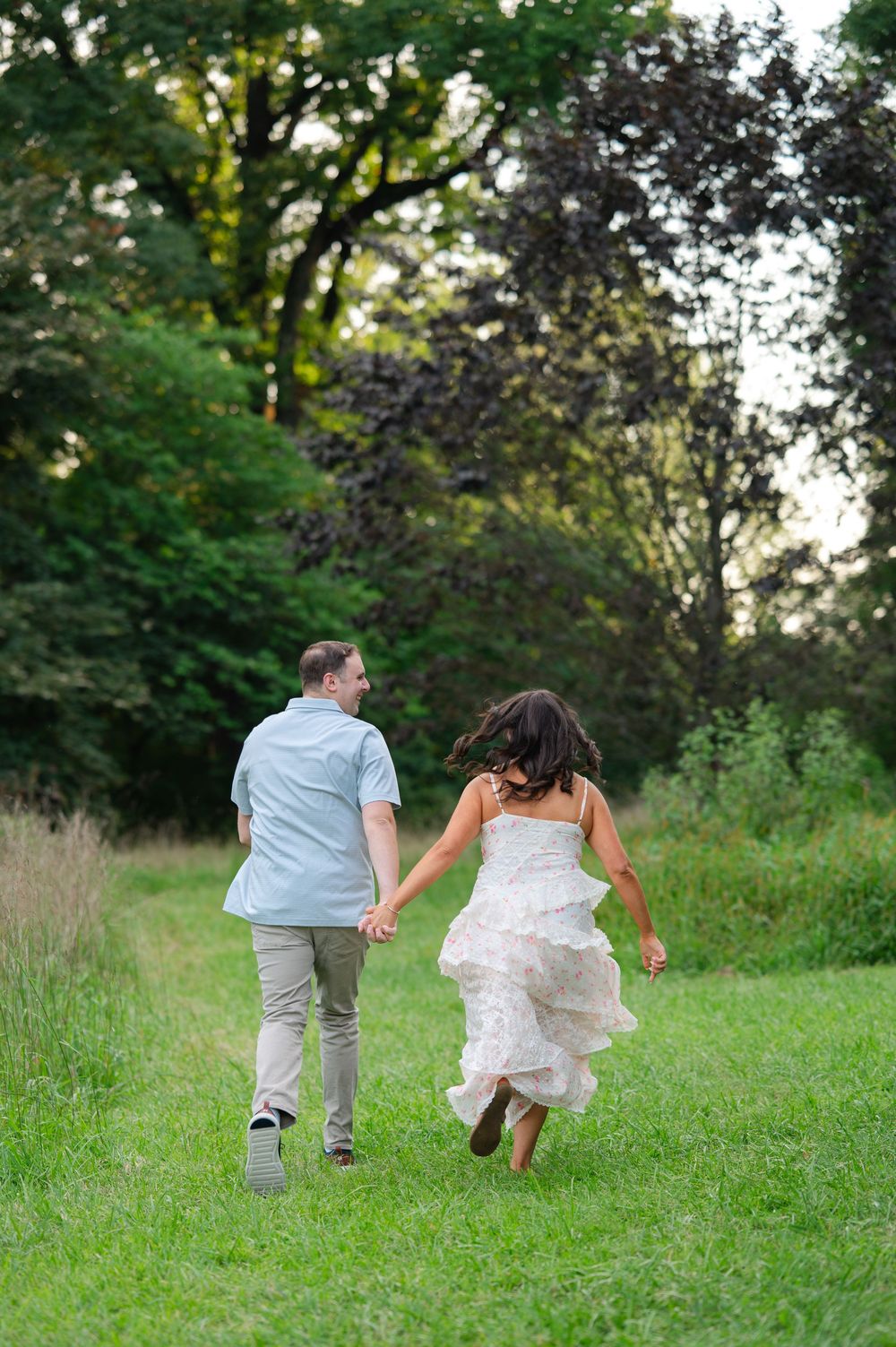 Couple in wedding attire holding hands running through lush green meadow toward trees.
