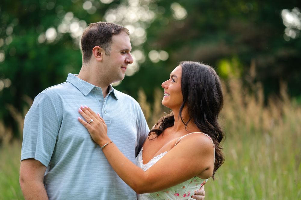 Couple smiling at each other in a sunlit field, woman in floral dress with hand on mans chest during engagement photo.