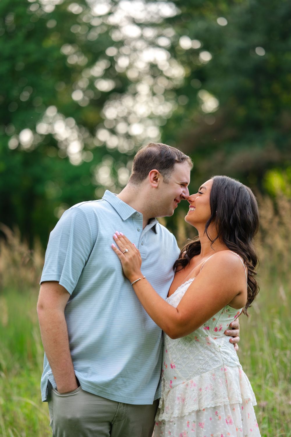 Smiling engaged couple touching noses in a meadow, woman wearing floral dress with engagement ring visible.