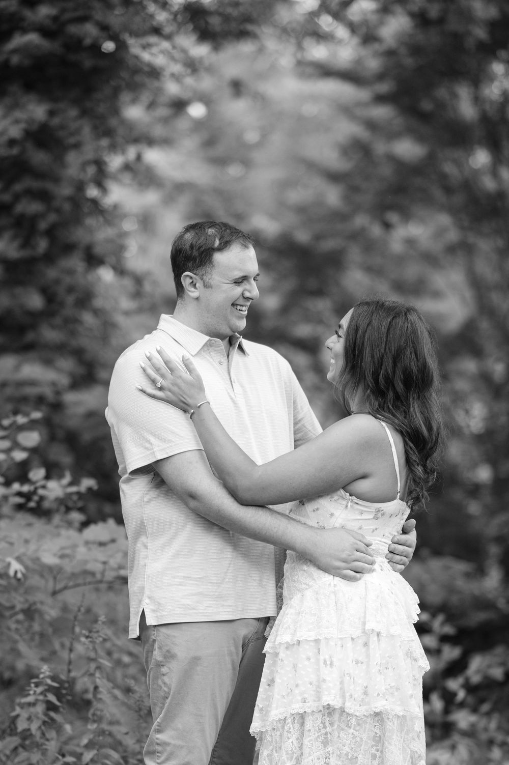 Black and white photo of a smiling couple embracing outdoors among lush green trees.
