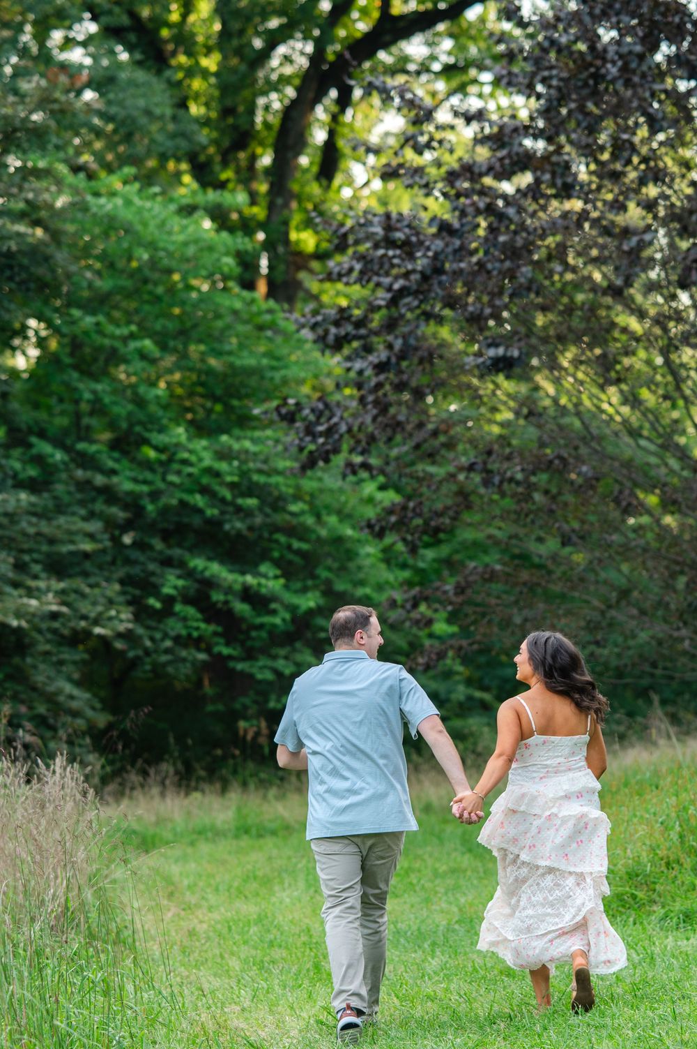 A couple holds hands walking through a lush green meadow toward trees, woman in white dress, man in blue shirt.