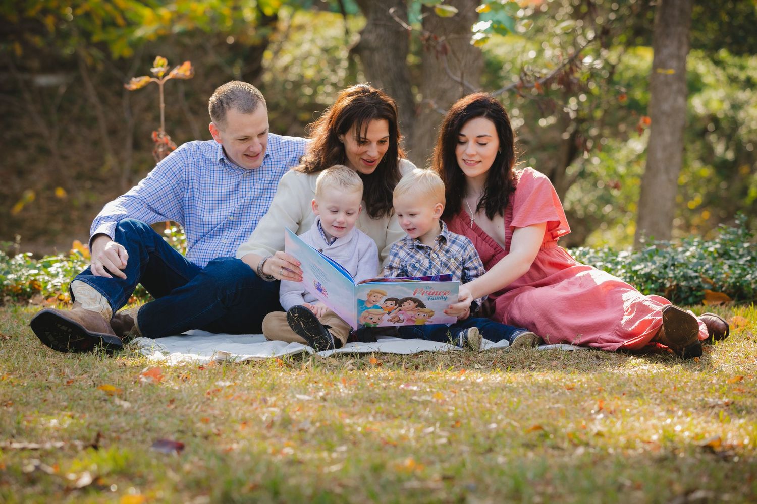 Family of five sitting outdoors on blanket reading a book together in autumn park setting.
