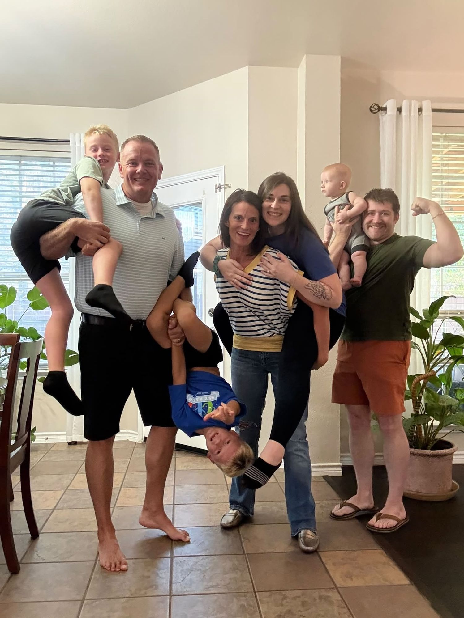 A joyful family group photo indoors with adults holding children on shoulders and upside down, all smiling and flexing.