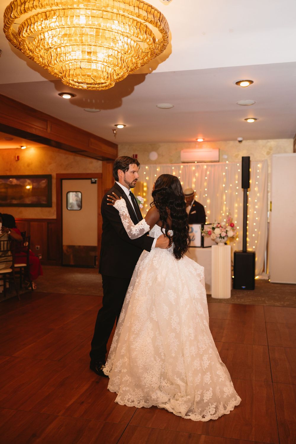 Interracial couple shares first dance under golden chandelier in warmly lit wedding reception venue.