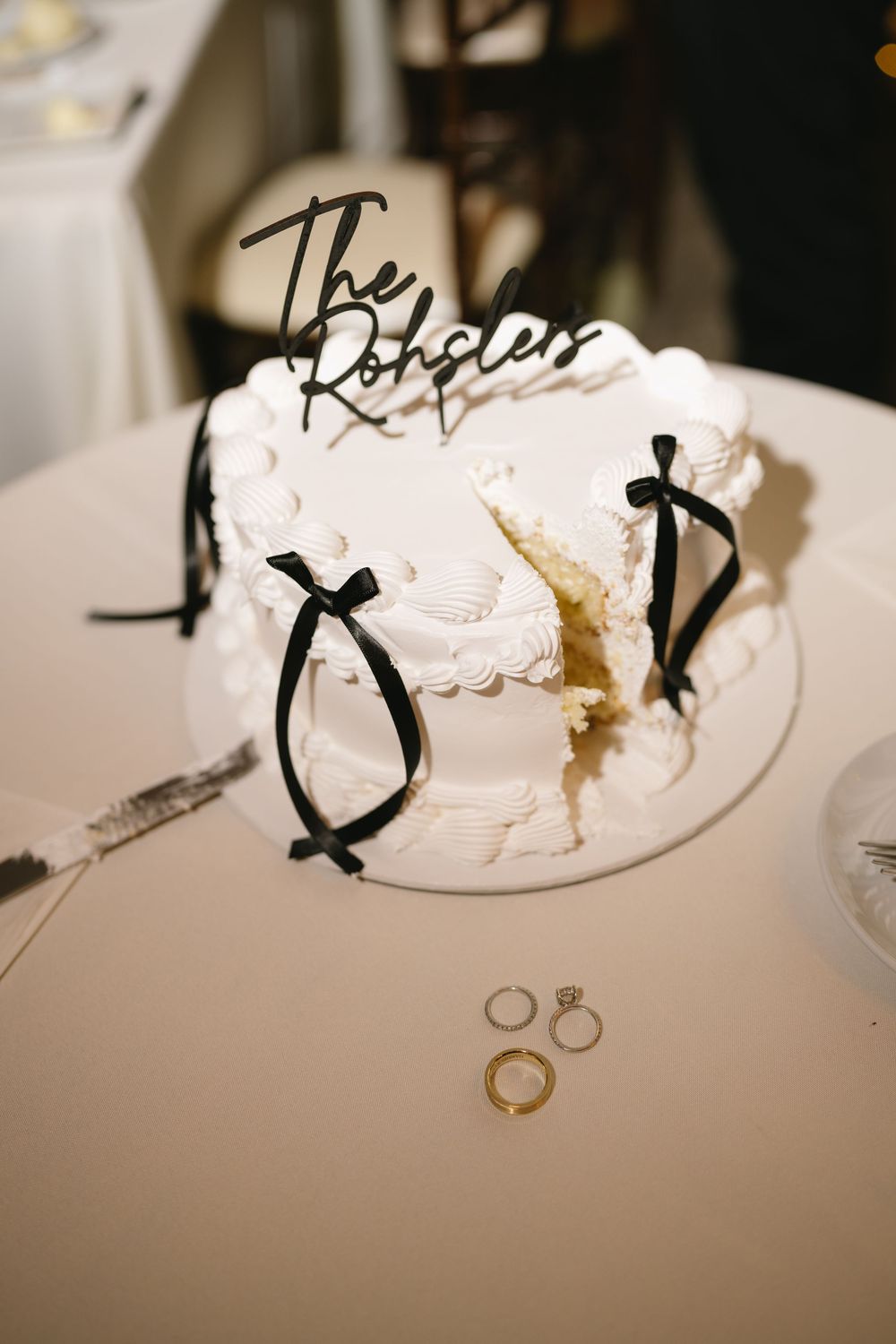 White wedding cake with black bow accents and The Rohslers topper displayed beside gold wedding rings.