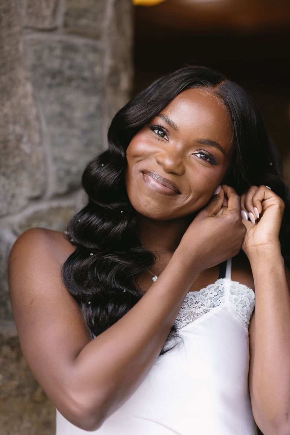 Beautiful Black bride smiles warmly while adjusting earring, wearing white lace bridal lingerie indoors.
