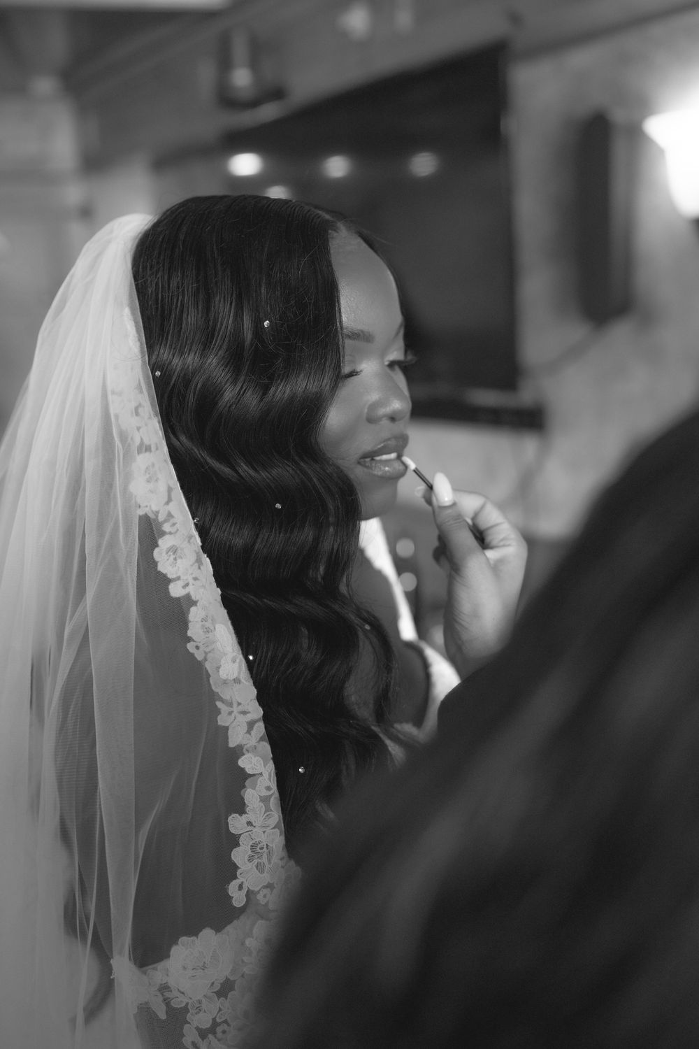 Black and white photo of a bride having lipstick applied while wearing a lace veil on her wedding day.