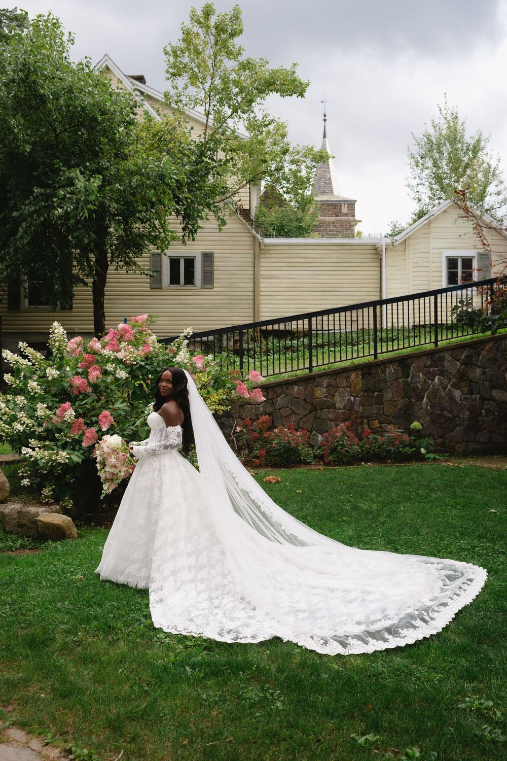 Bride in a lace ball gown with cathedral veil poses beside blooming flowers in a lush garden setting.