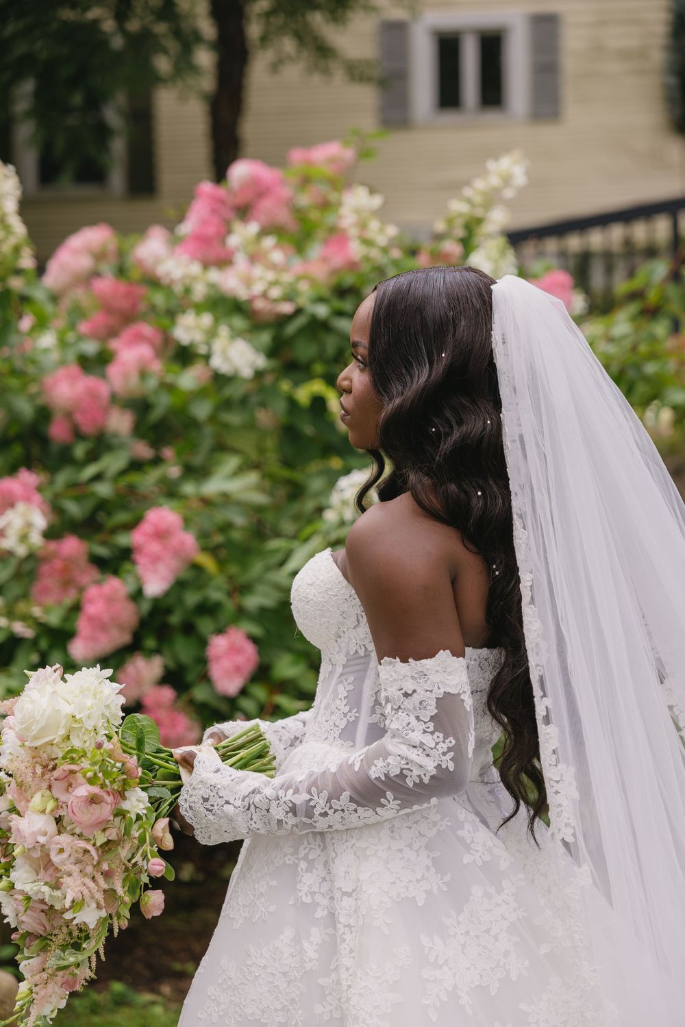 Bride in lace gown and veil stands near pink hydrangeas holding a white bouquet, back view.