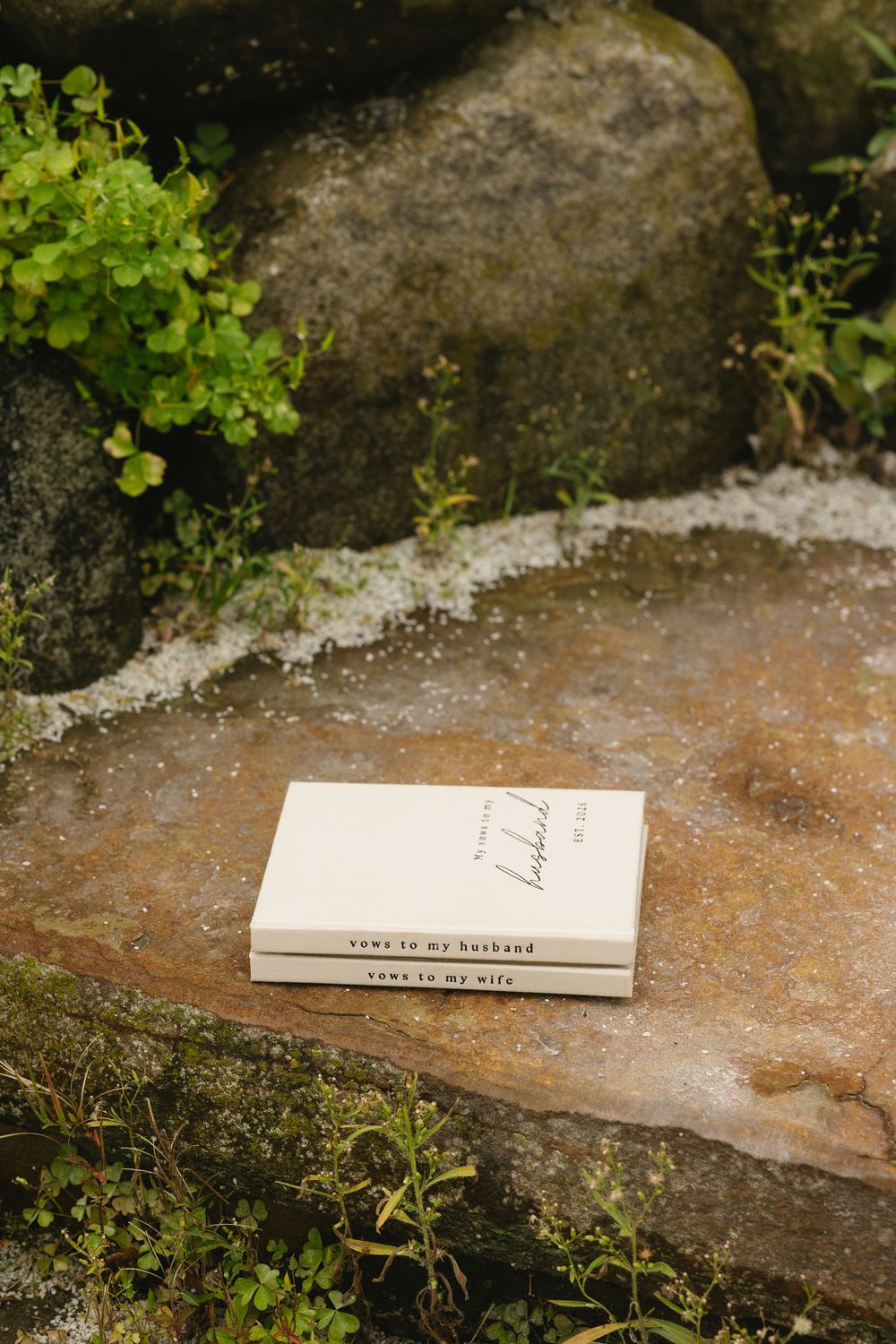 Two wedding vow books labeled vows to my husband and vows to my wife resting on a mossy stone surface.