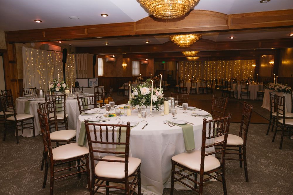 Elegant wedding reception hall with round tables, chiavari chairs, white linens, floral centerpieces, and warm string light backdrop.