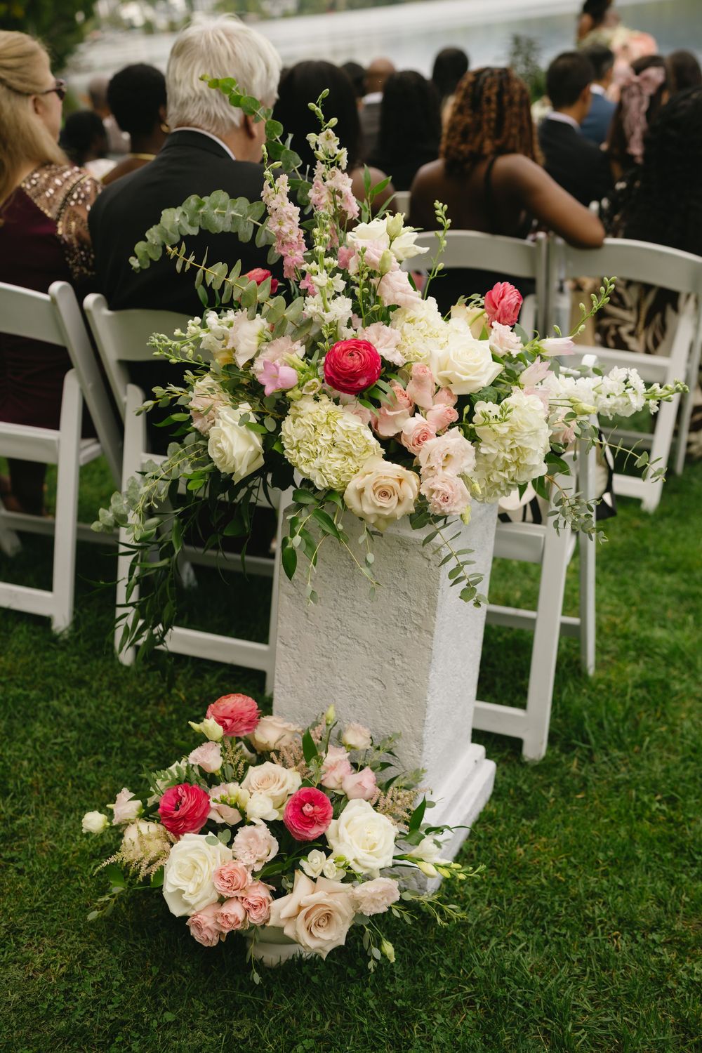 Elegant wedding ceremony aisle with white pedestal floral arrangements featuring pink roses, hydrangeas, and greenery on green grass.