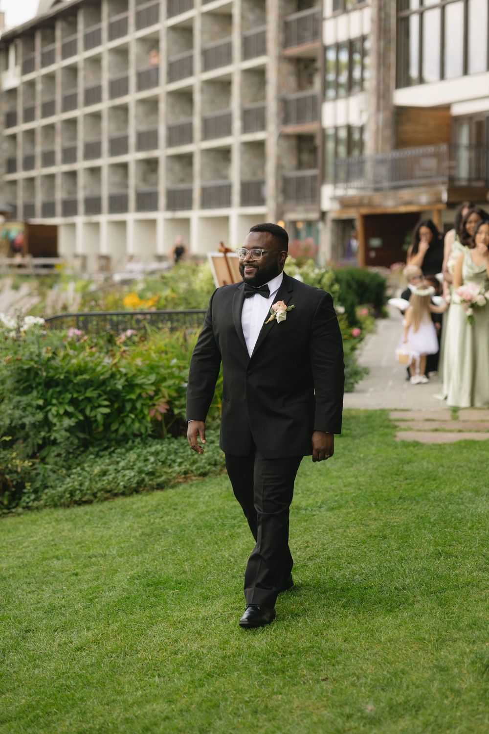 Groom in black tuxedo with bow tie and boutonniere walks across green lawn at outdoor wedding venue.