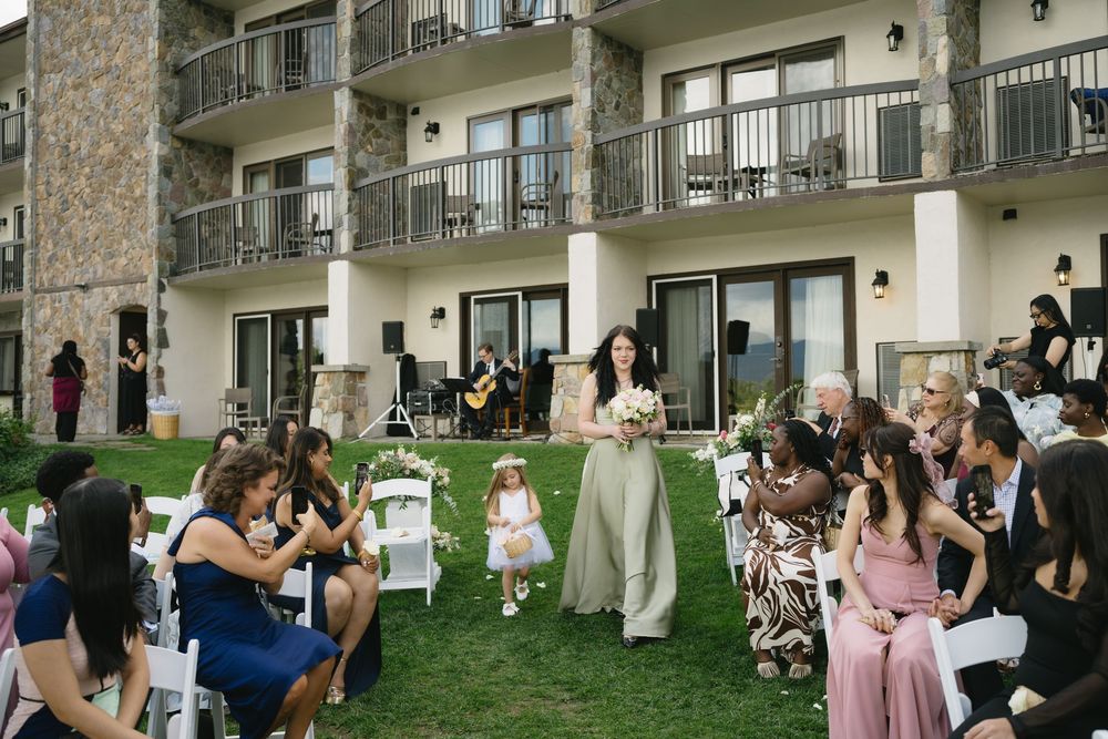 A bridesmaid and flower girl walk down the aisle at an outdoor wedding ceremony beside a stone hotel.
