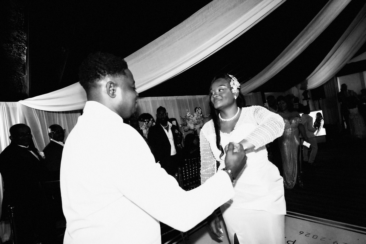 Black and white photo of a groom in a white suit holding hands with a smiling bride in white during a wedding reception dance.