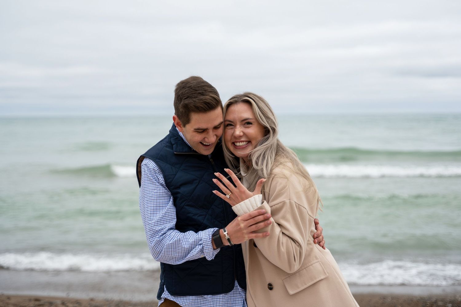 Smiling couple embracing on a beach as the woman shows off her new engagement ring, with gentle waves and an overcast sky behind them.