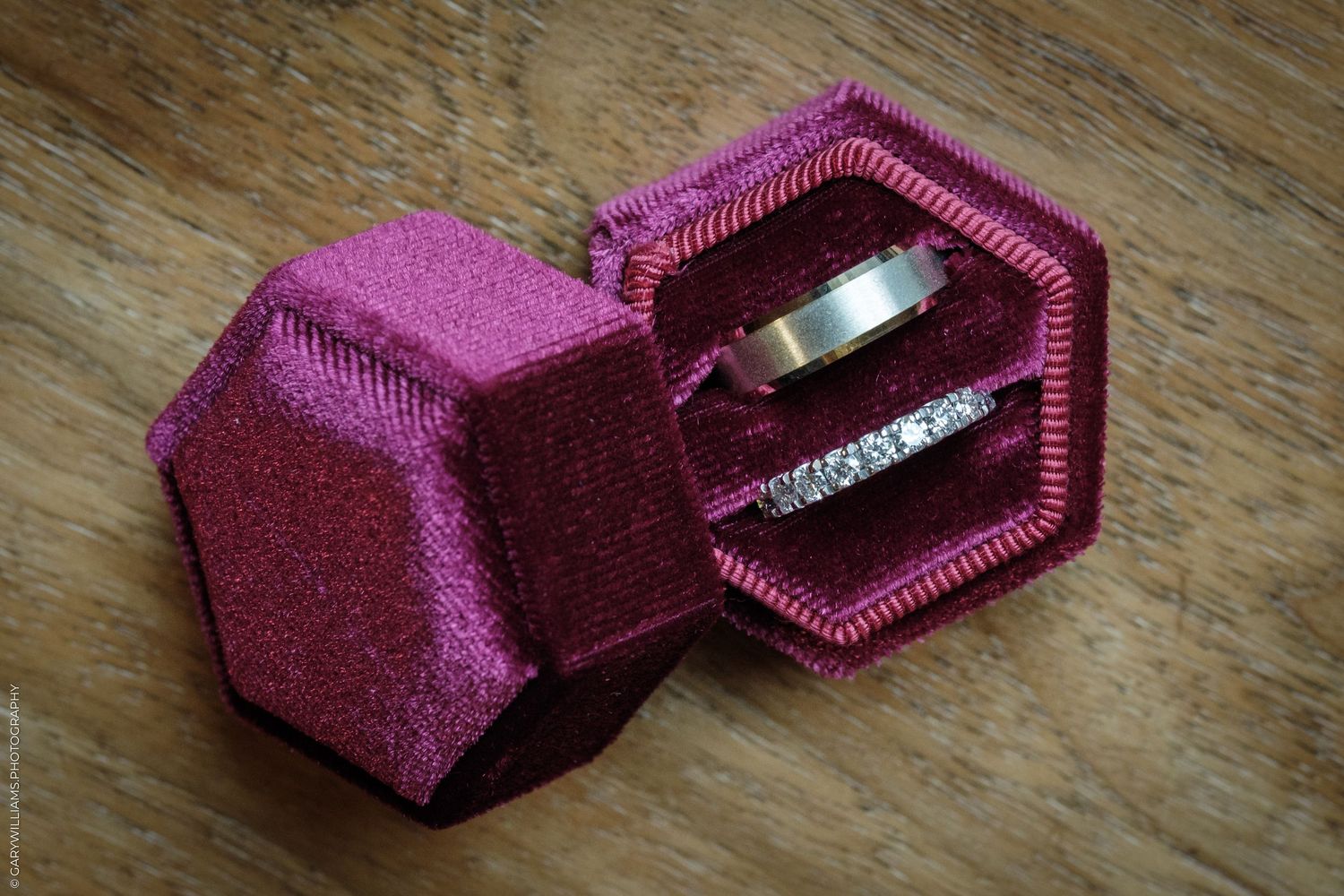 Open burgundy velvet hexagonal ring box holding a plain metal wedding band and a diamond-studded band on a rustic wooden surface.