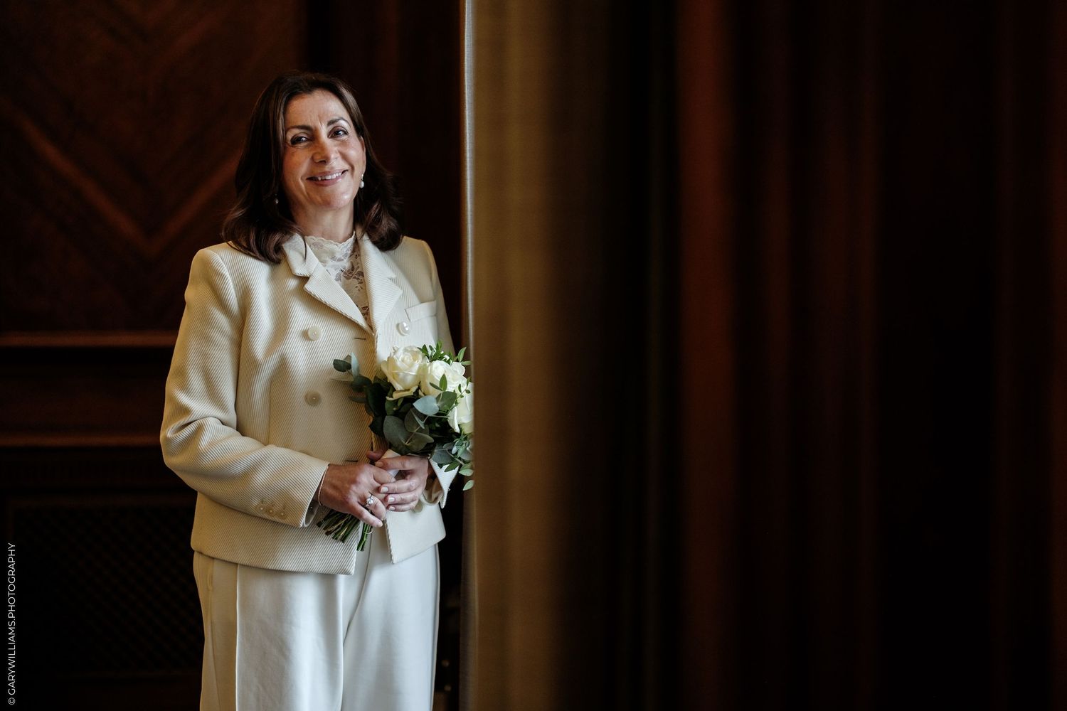 Smiling woman in a cream tweed jacket and white trousers holds a small bouquet of white flowers beside dark wood paneling and a curtain.