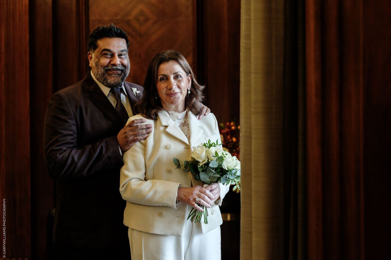 Smiling groom in a dark suit stands beside his bride in a cream jacket holding a white bouquet at an elegant wedding ceremony.