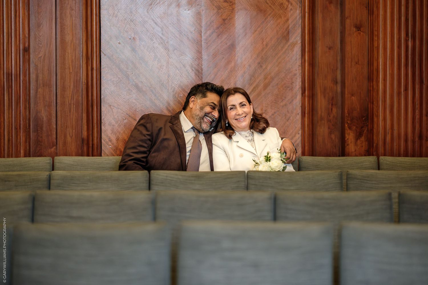 Smiling couple in formal attire sit close together, leaning heads, on wooden courtroom benches with warm paneled walls behind them.