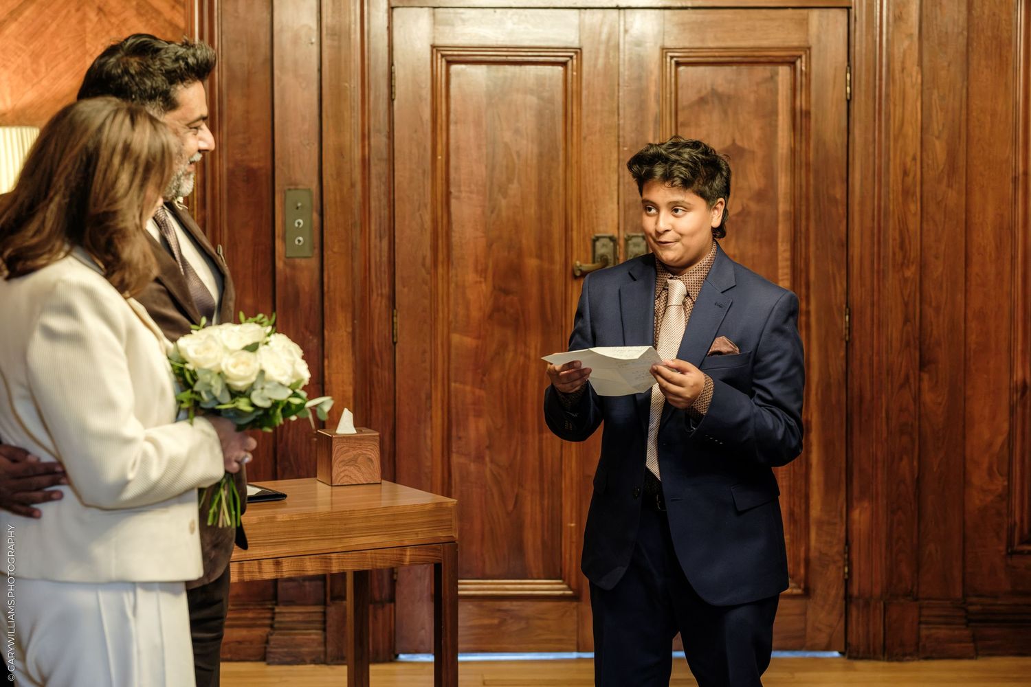 A young person in a navy suit reads from a paper during an intimate wedding ceremony while a bride holding a white bouquet stands nearby.