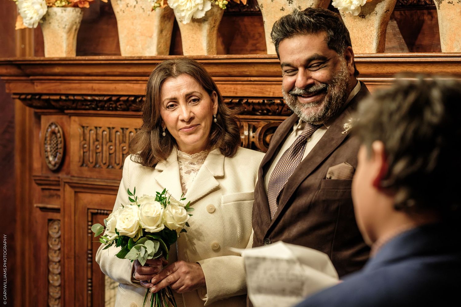 Smiling bride in a cream coat holding a white rose bouquet stands beside a bearded groom in a brown suit during a wedding ceremony.