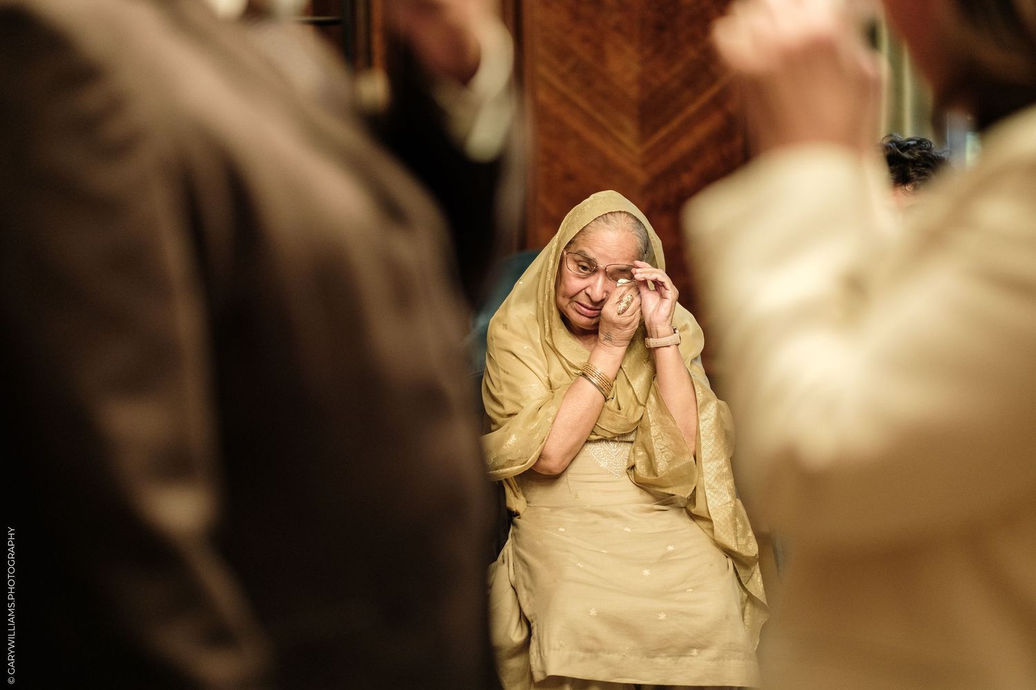 Elderly woman in a cream headscarf and embroidered outfit wipes tears from her eyes while standing between blurred guests at an event.