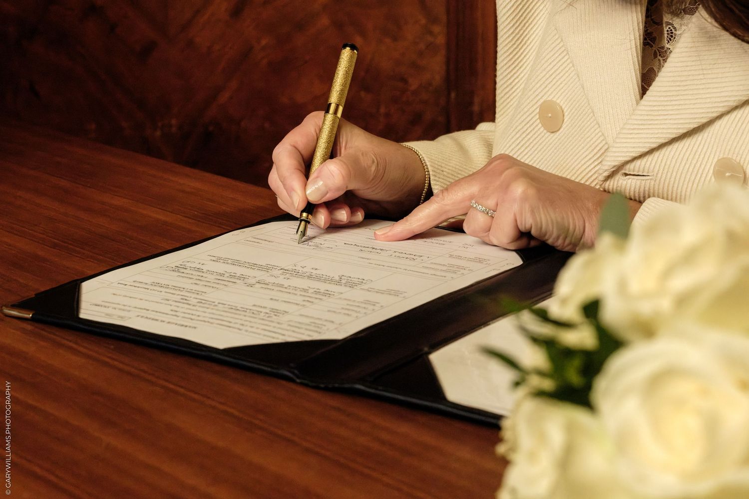 Woman in a cream blazer signs a document with a gold pen on a polished wooden table beside white roses.