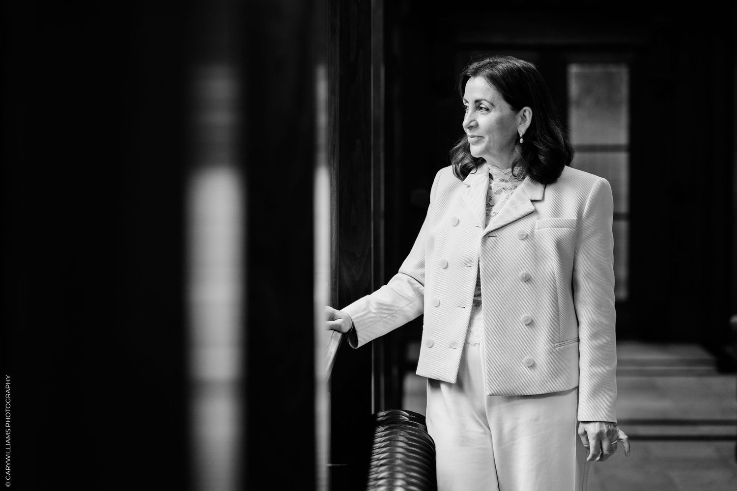 Black and white portrait of a smiling woman in a light double-breasted blazer standing in a dark hallway.