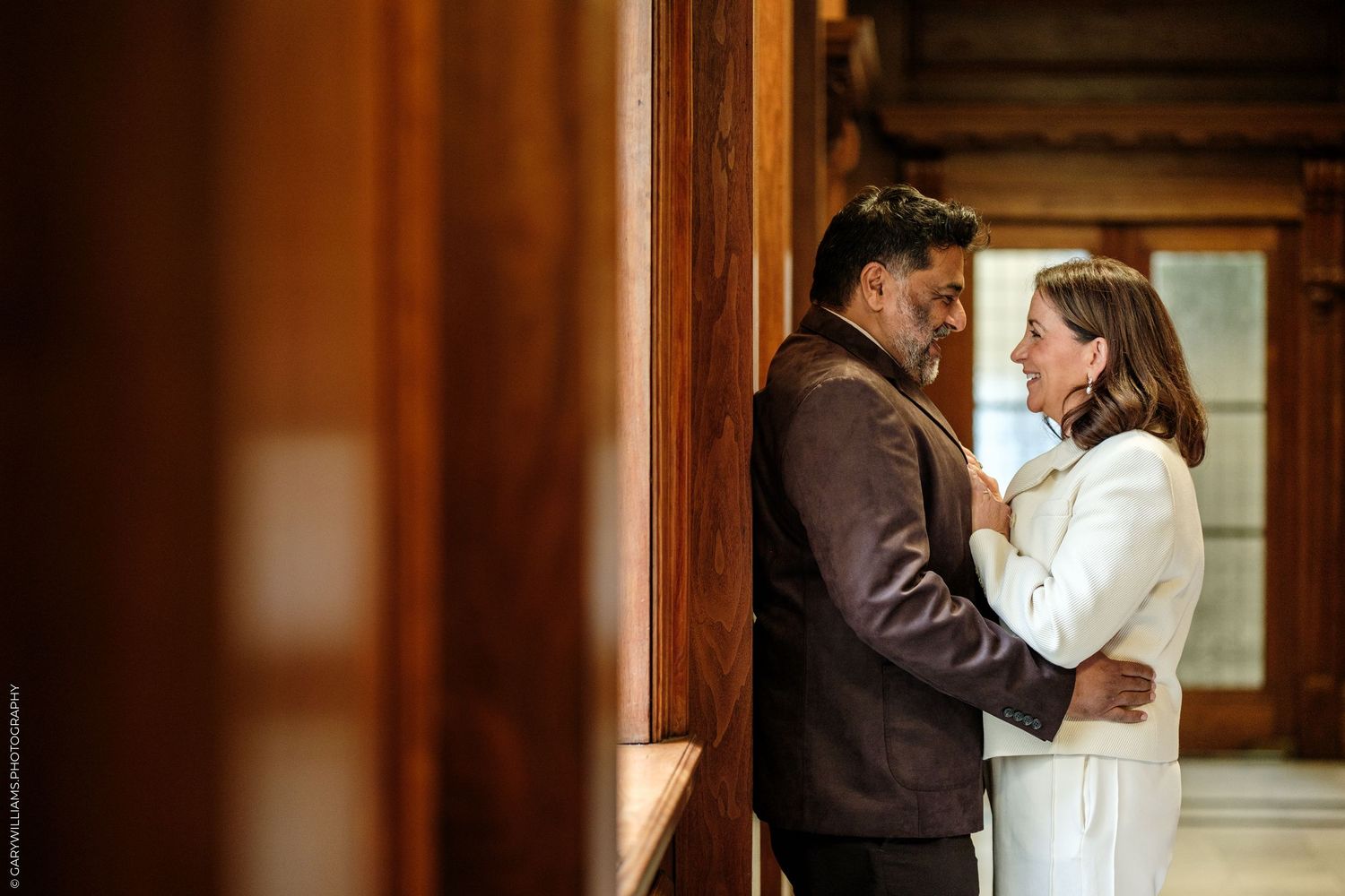 A couple in an affectionate embrace beside a wood-paneled window, the woman in a white outfit and the man in a dark brown jacket.