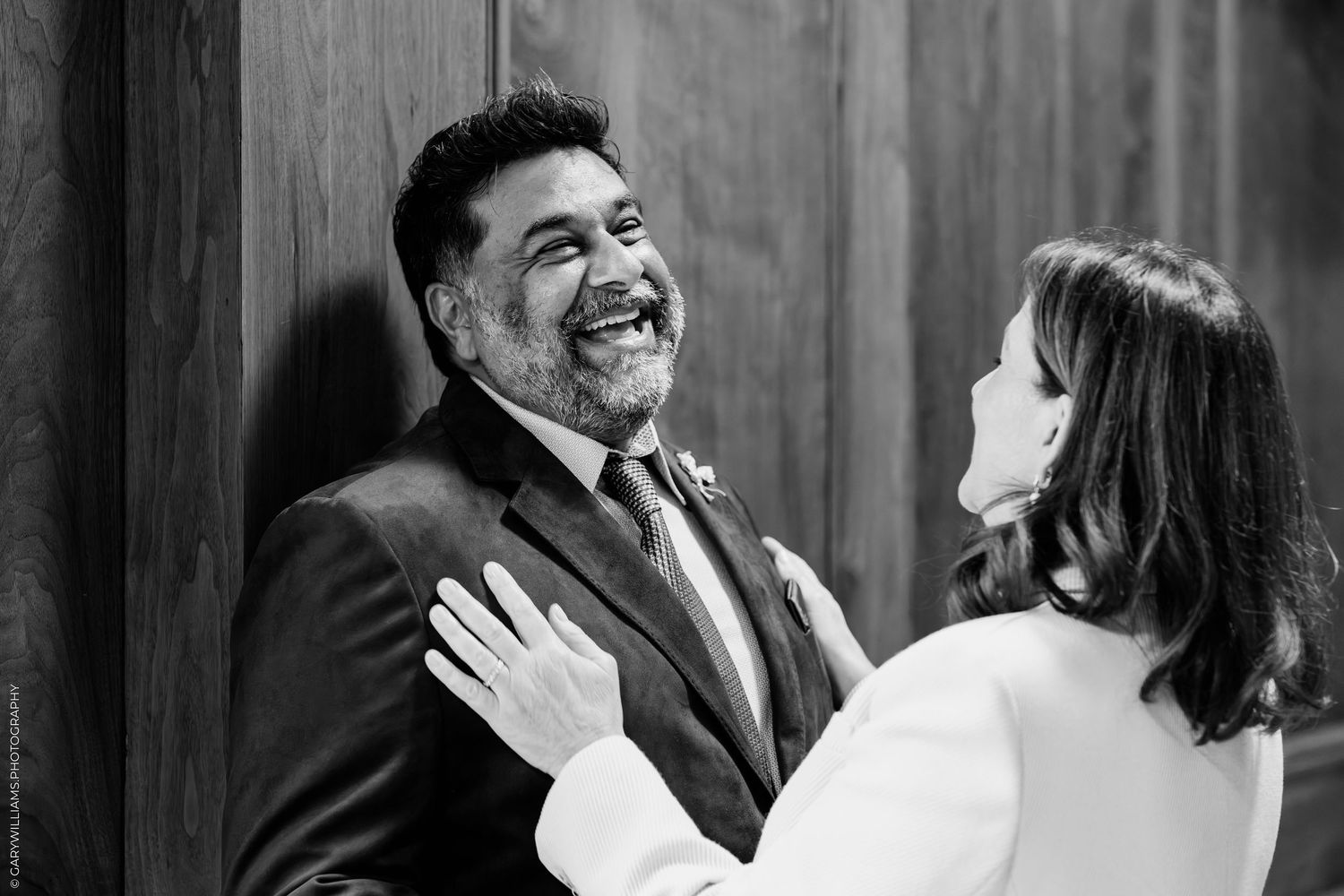 Black and white photo of a smiling bearded man in a suit laughing as a woman in a white jacket rests her hand on his shoulder.