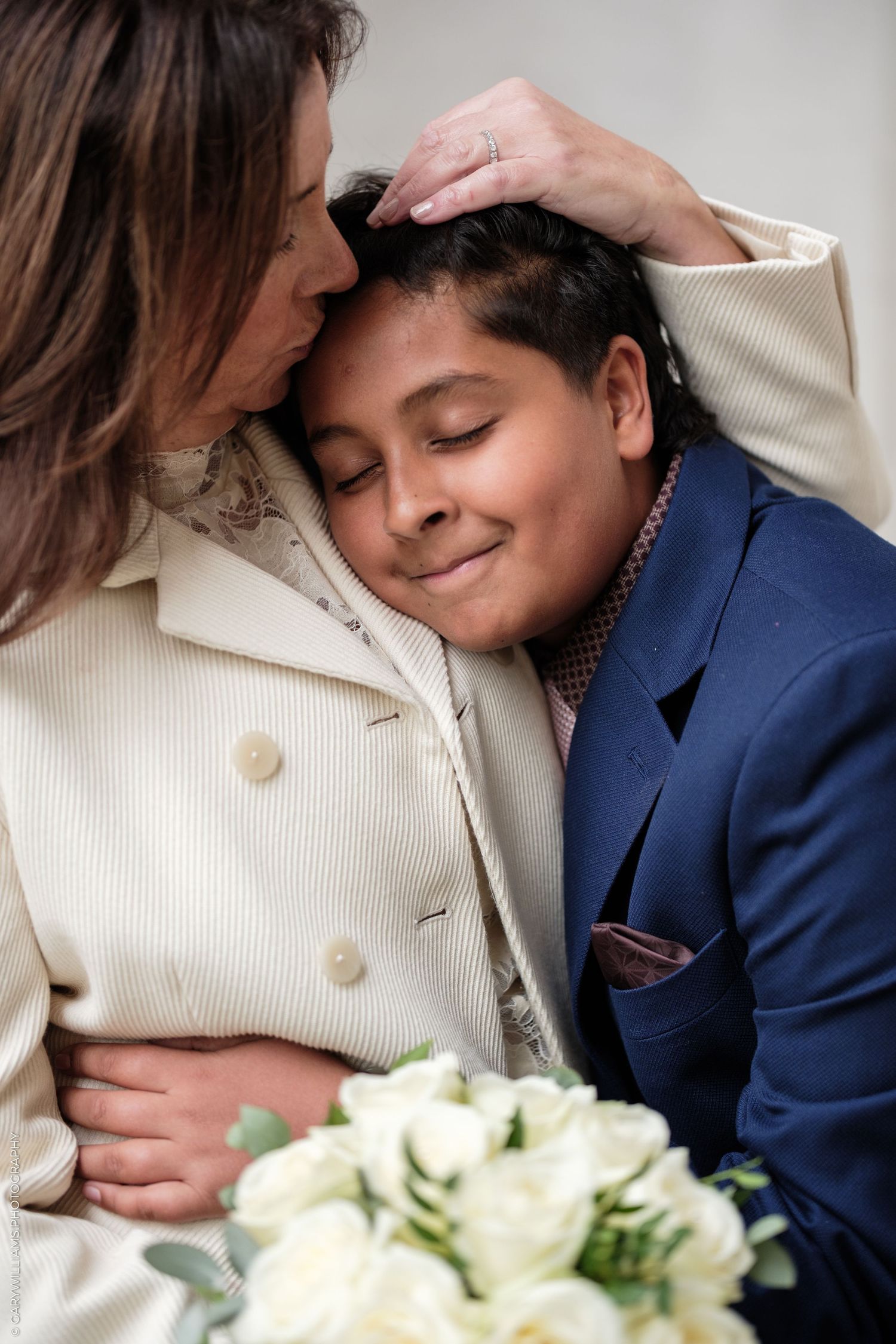 A woman in a cream jacket tenderly kisses the forehead of a smiling boy in a navy suit, with white roses in the foreground.