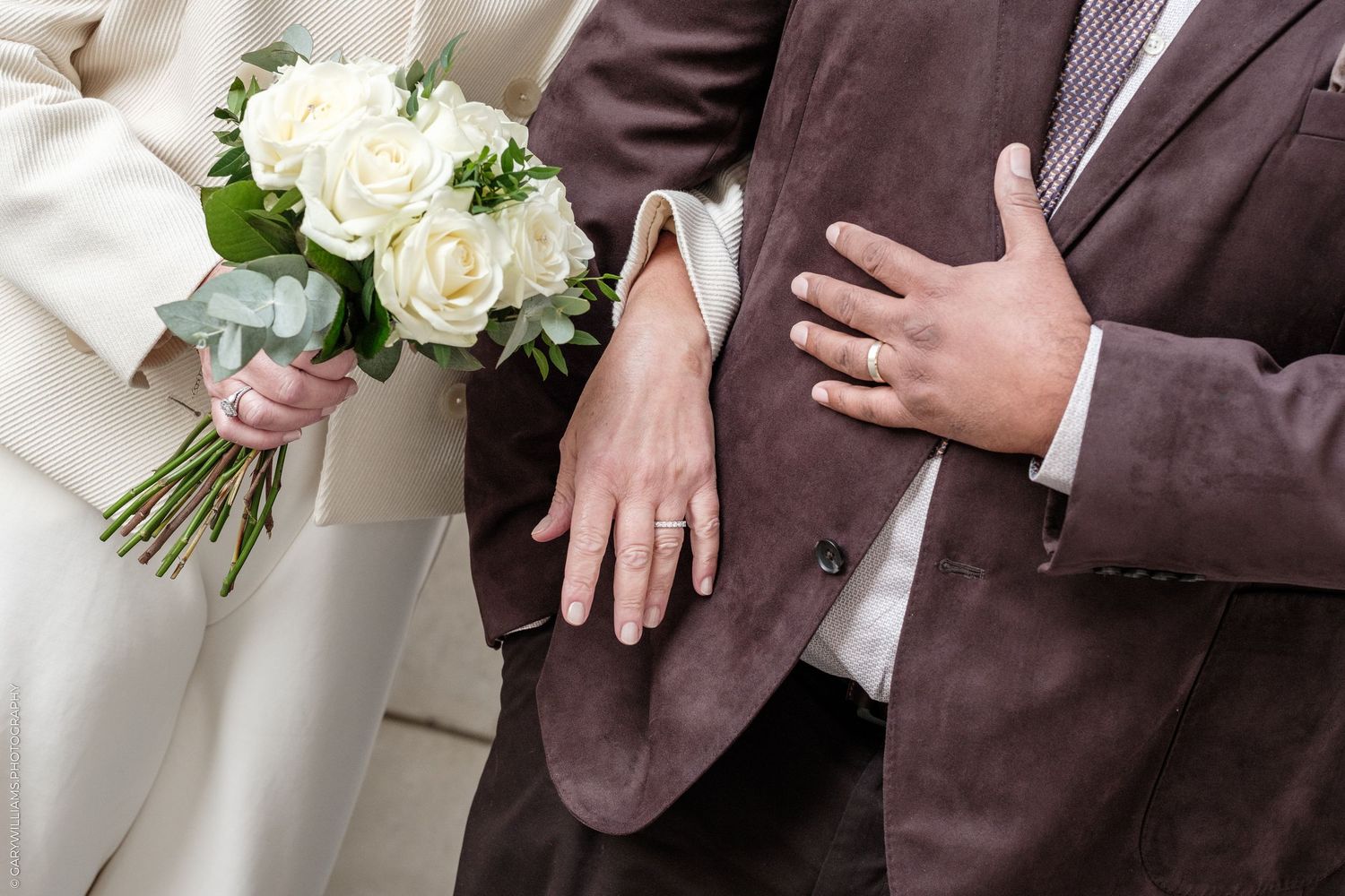Close-up of a bride holding a white rose bouquet as the groom in a brown suit rests his hand on hers, both wearing wedding rings.