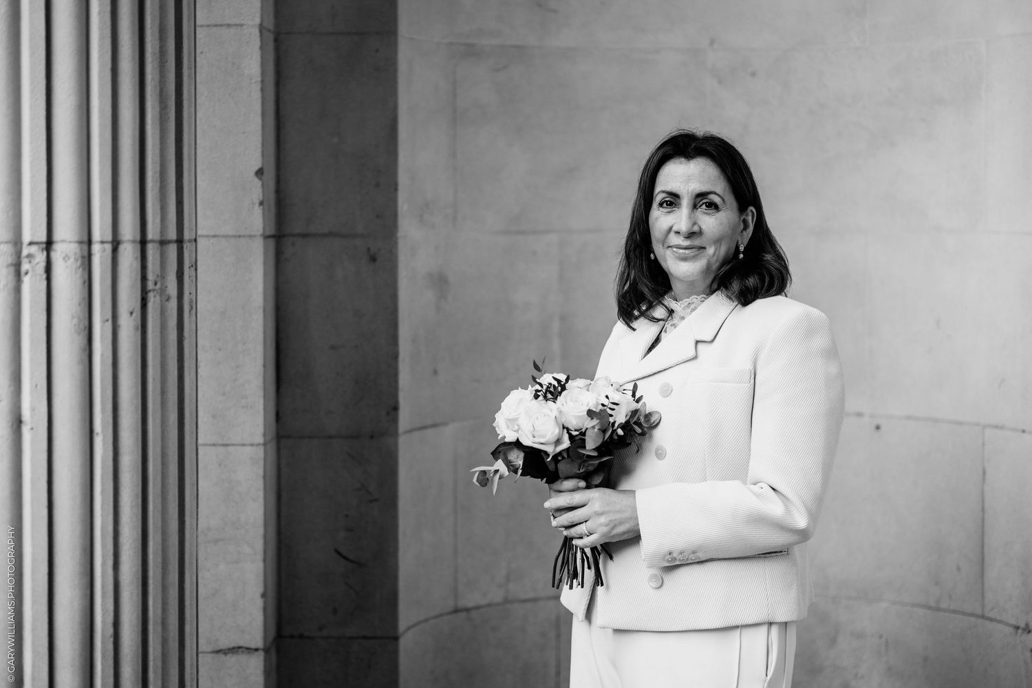 Black and white portrait of a smiling bride in a light suit holding a bouquet of flowers beside stone columns.