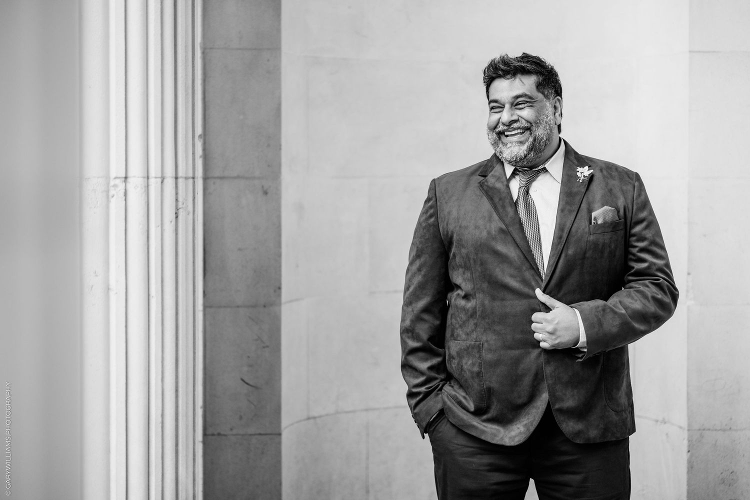 Smiling bearded man in a tweed suit and tie poses with one hand in his pocket against a stone building, in black and white.