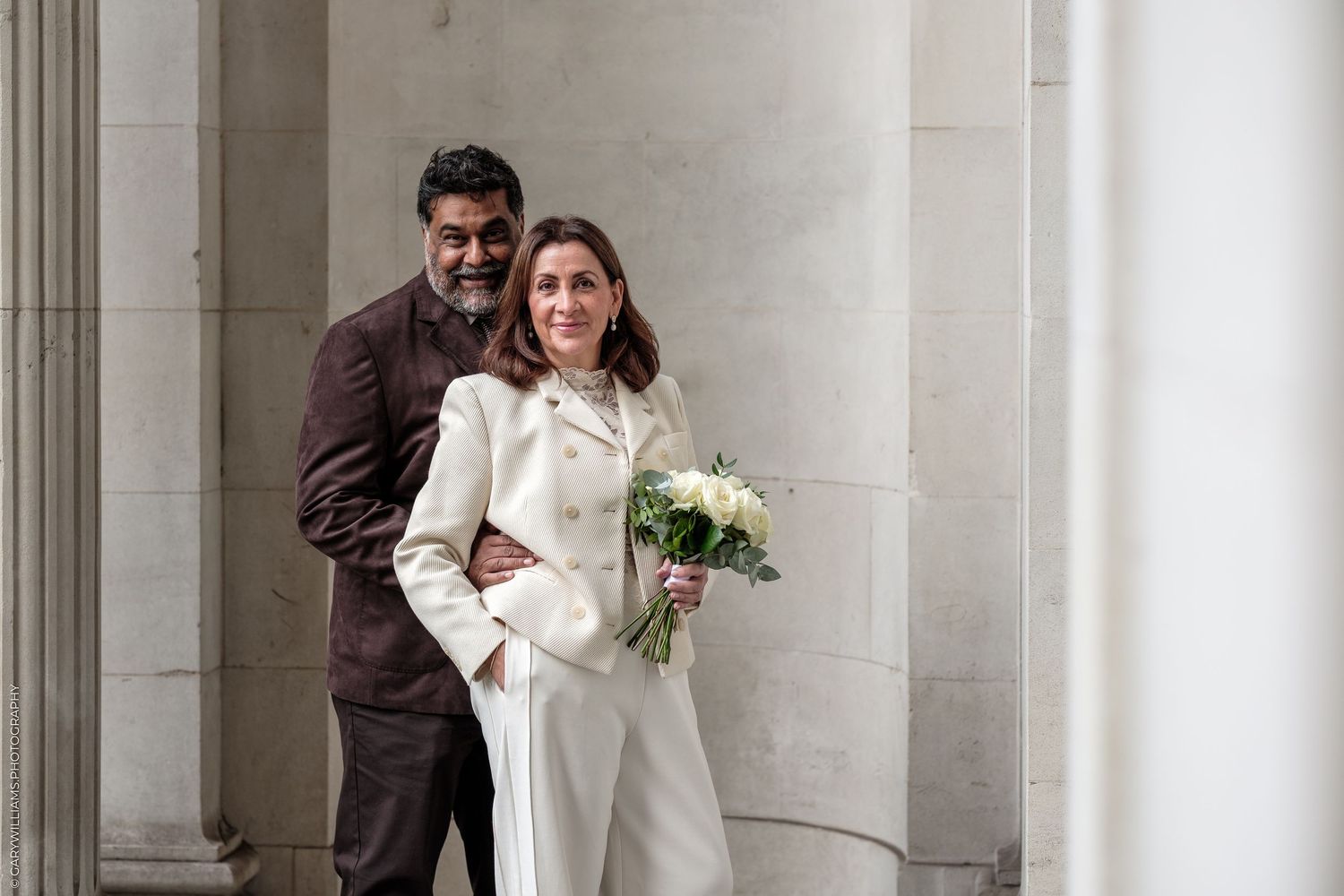 Smiling bride in a white suit holding a bouquet of white flowers while her groom embraces her between stone columns.
