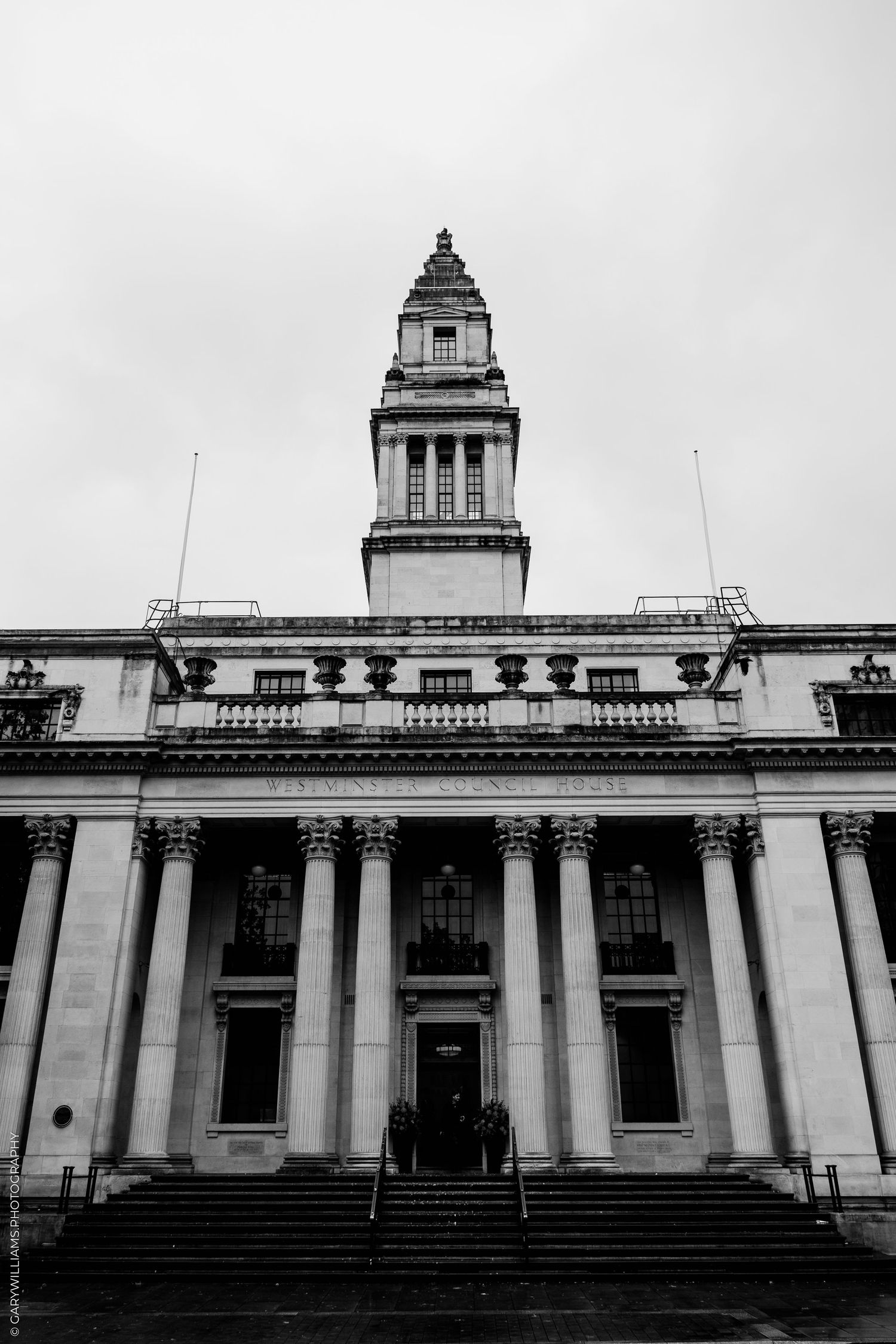 Black and white photo of a neoclassical building with tall columns, wide front steps and a clock tower rising against an overcast sky.