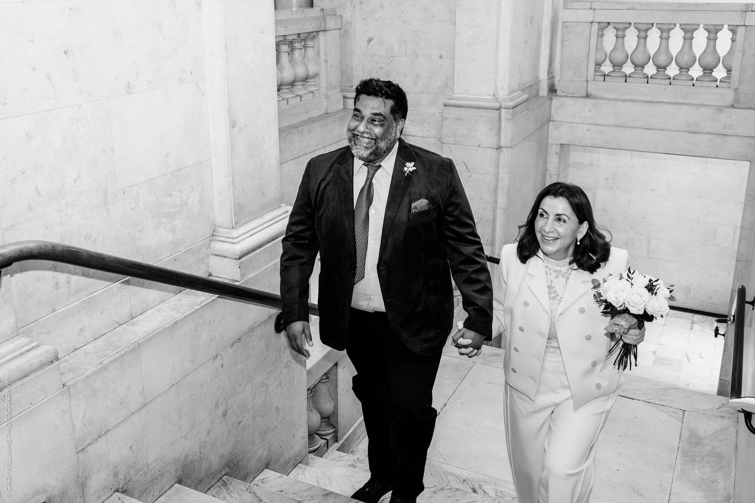 Smiling groom in a dark suit holds hands with a bride in a white jacket carrying a bouquet as they climb stone stairs in black and white.