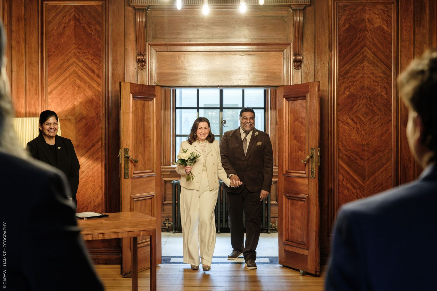 Bride in white pantsuit holding a bouquet walks hand in hand with groom through wood-paneled doors at a civil wedding ceremony.