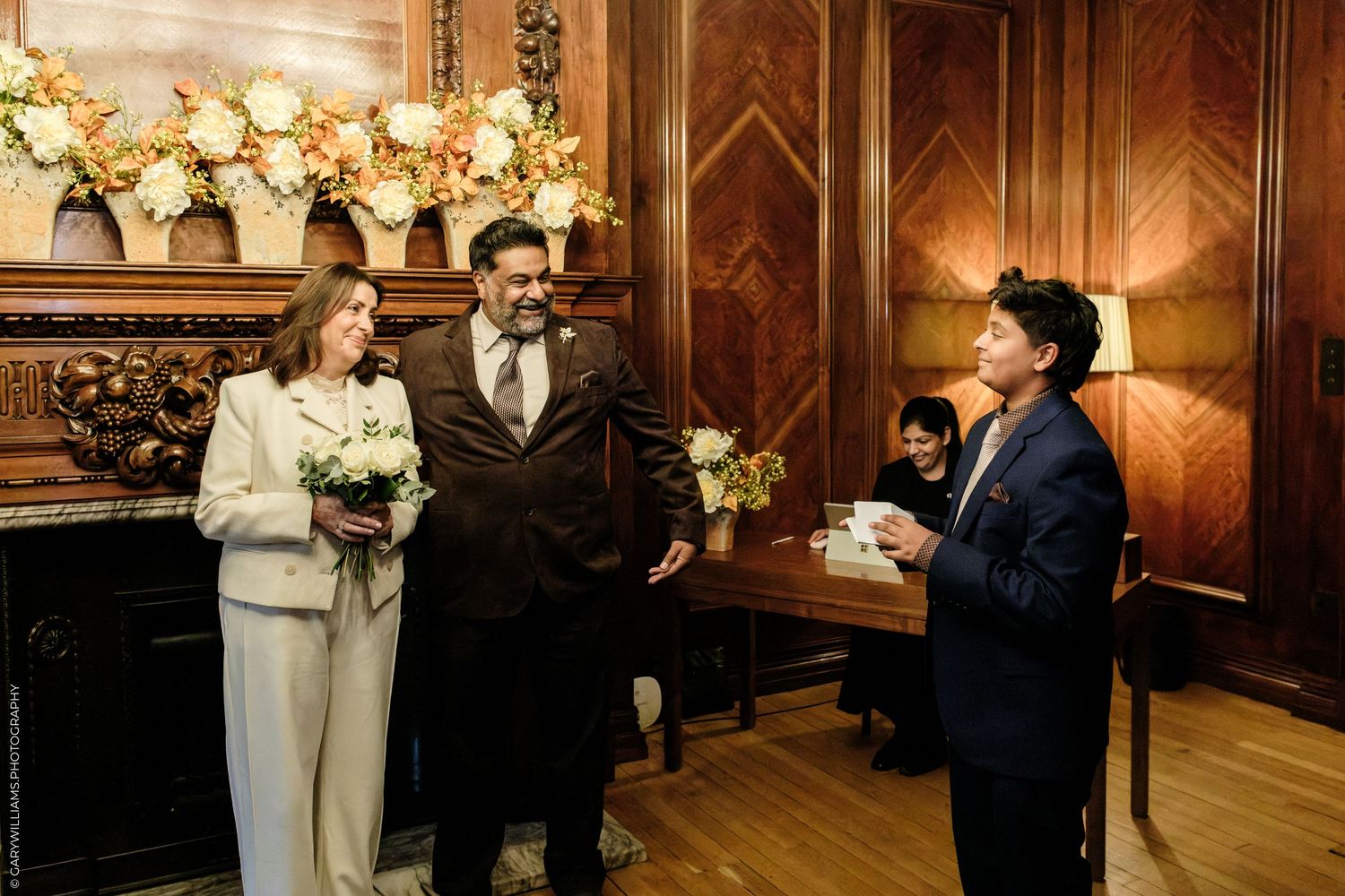 A couple stands in a wood-paneled room with floral arrangements as an officiant reads a document during an intimate civil wedding ceremony.