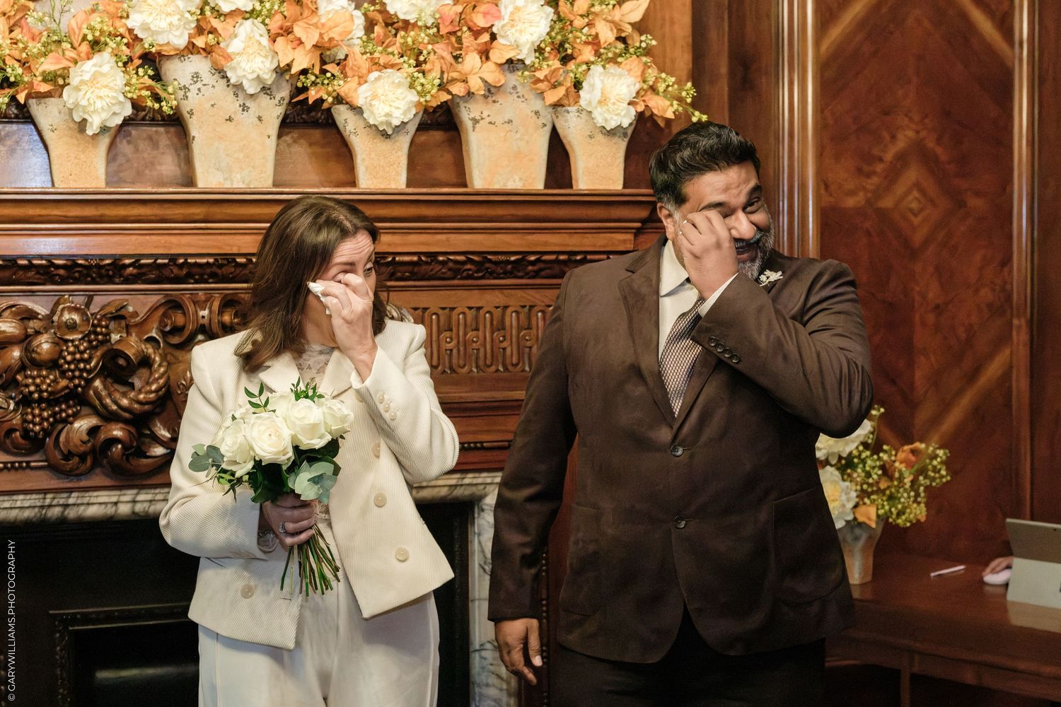 Emotional bride in a white suit holding a white bouquet and groom in a brown suit wiping tears during a ceremony in a wood-paneled room.