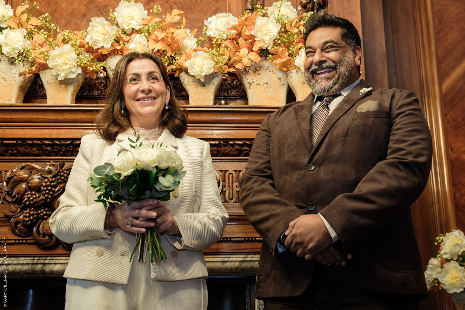 A smiling bride in a cream suit holds a white rose bouquet beside her groom in a brown jacket in an ornate wood-paneled room with flowers.