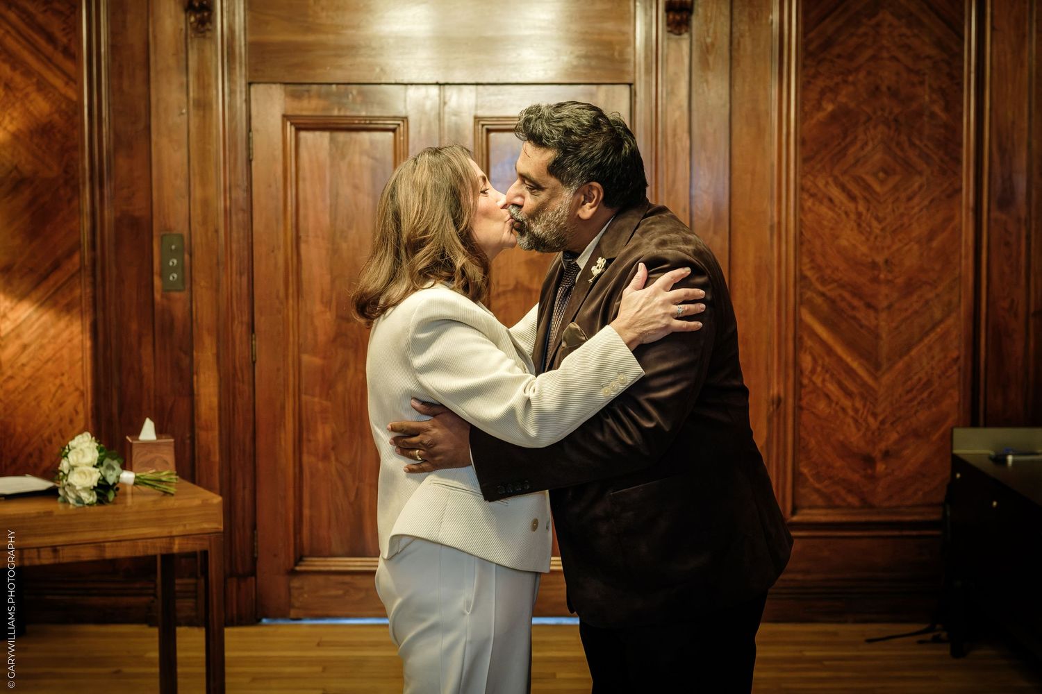 A woman in a white suit and a man in a dark jacket embrace in a wood-paneled room beside a small table with white flowers.