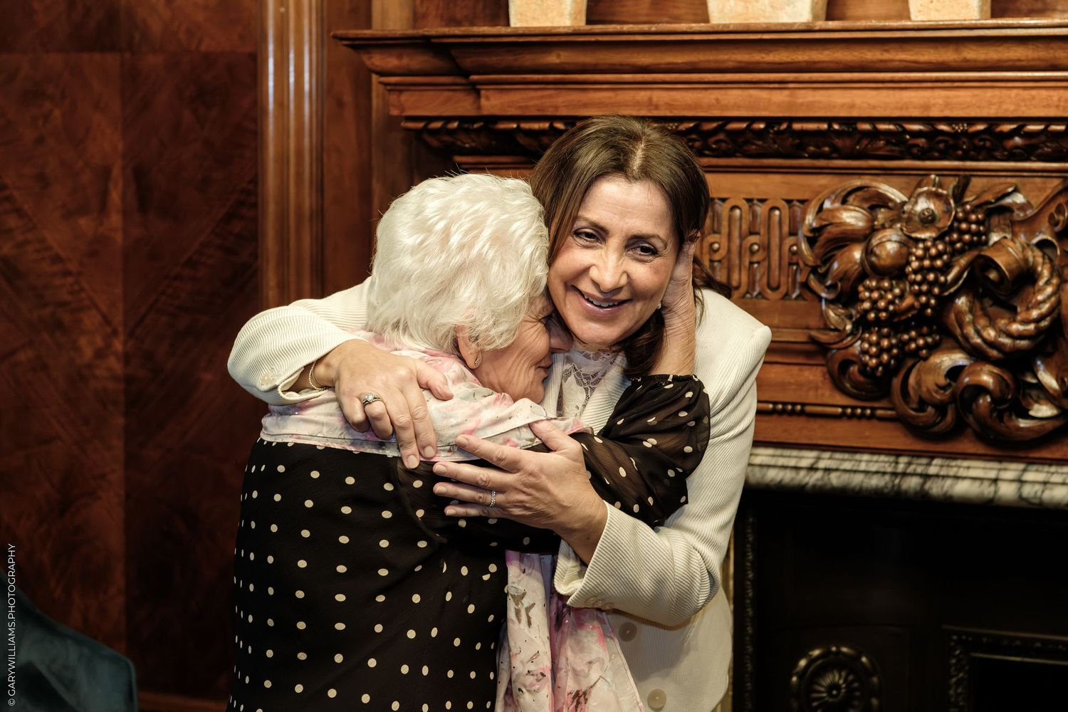 Two women share a warm embrace in a wood-paneled room with an ornately carved fireplace, one with white hair and one smiling.
