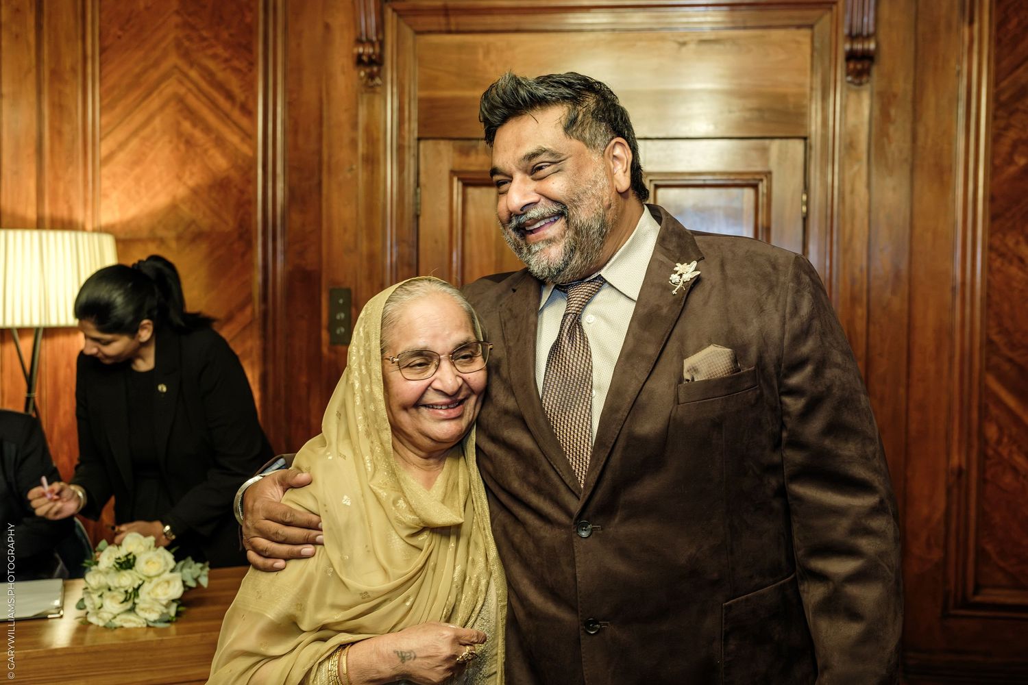 Smiling bearded man in a brown suit with a boutonniere embraces an elderly woman in a yellow headscarf in a wood-paneled room.