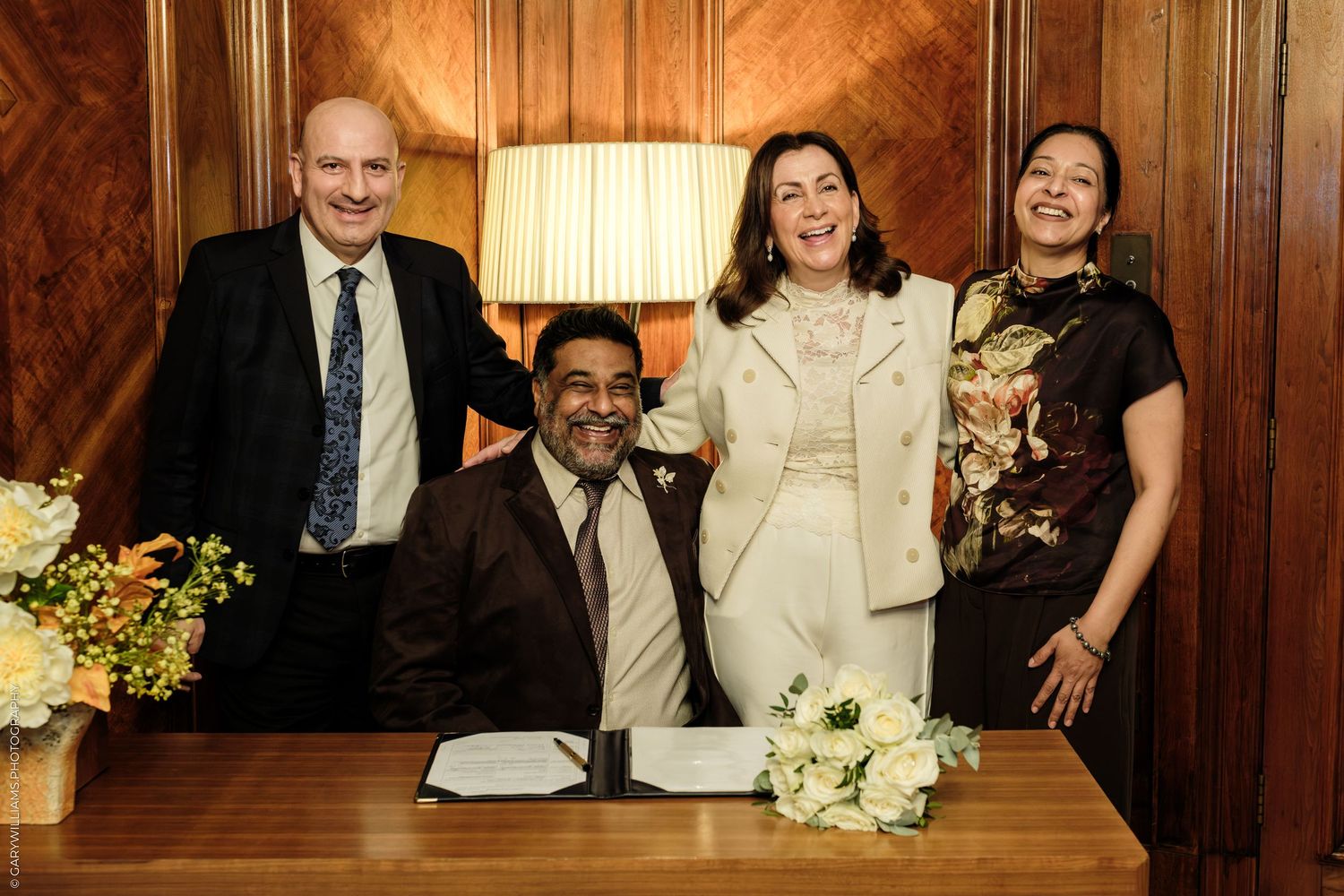 Smiling bride in white suit stands with three guests beside a signing table with bouquets in a wood-paneled civil ceremony room.