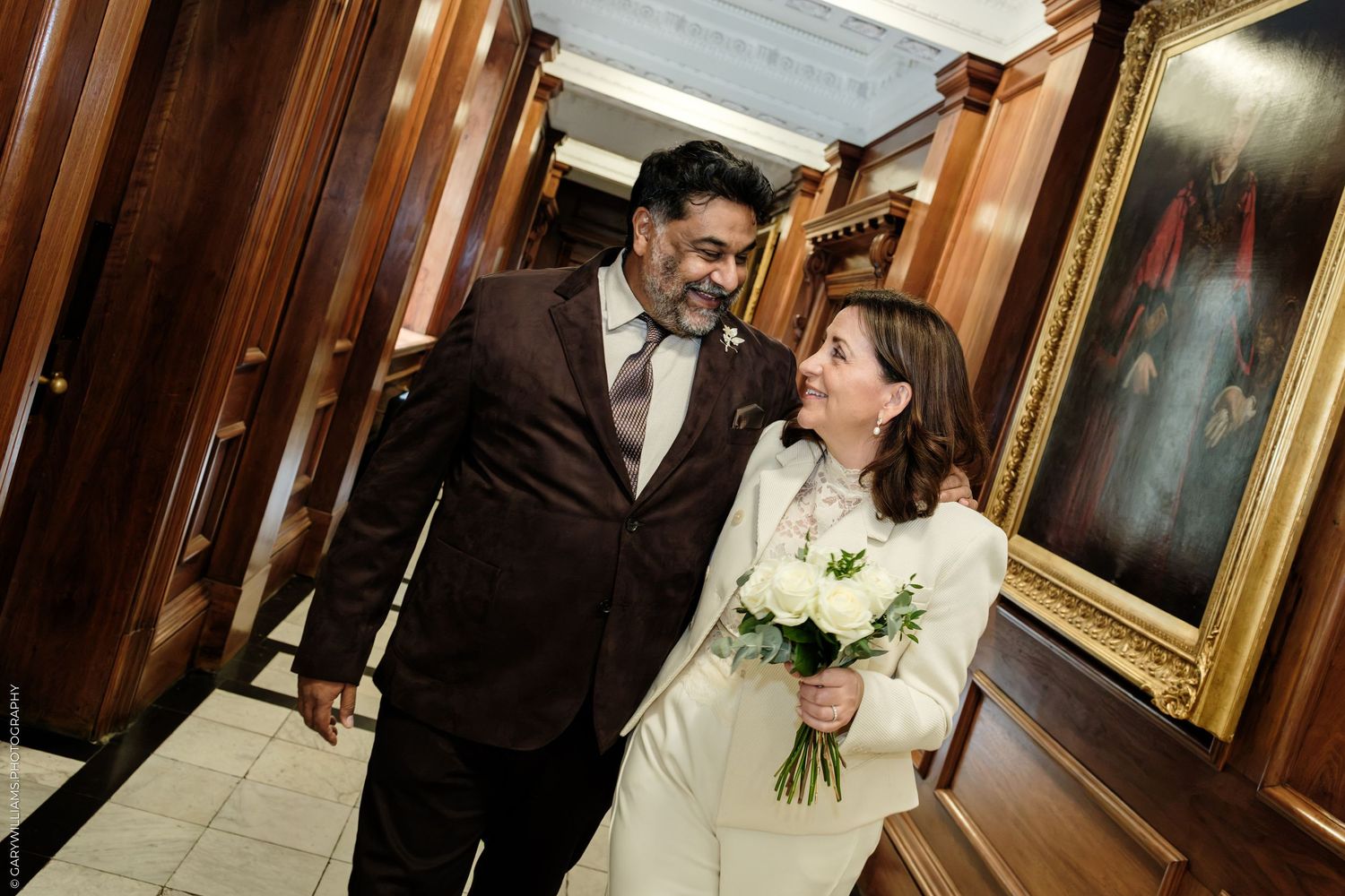 Smiling bride in a white suit holding a white floral bouquet walks beside her groom in a brown suit in a wood-paneled hall.
