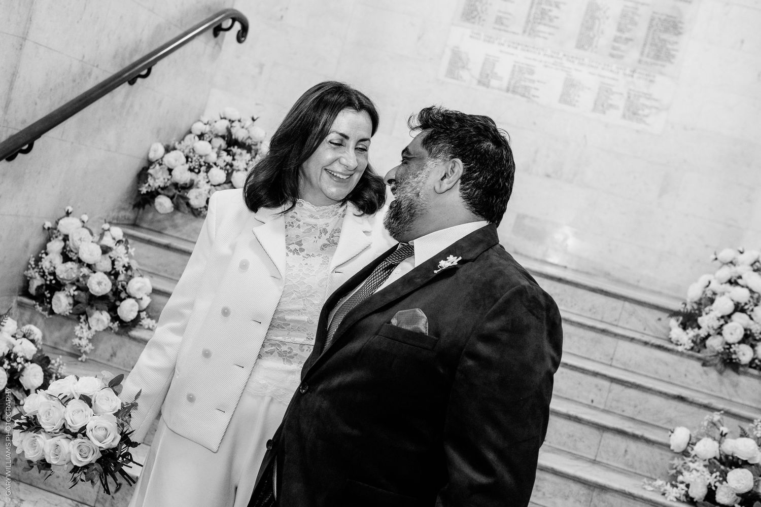 Black and white photo of a smiling couple in wedding attire on a staircase lined with white floral arrangements.
