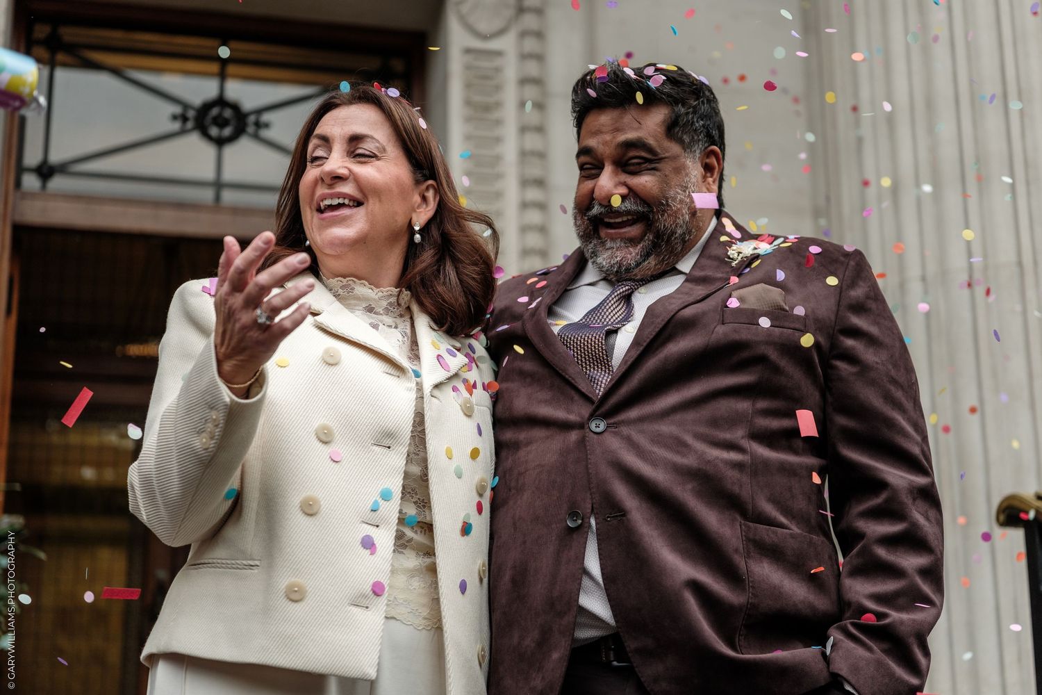 A smiling couple in light and brown jackets celebrates outdoors as colorful confetti falls around them in front of a building.